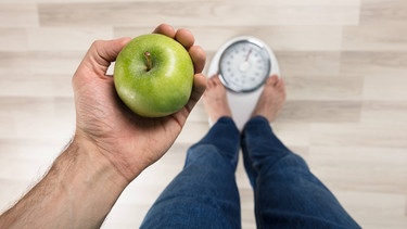 Ein Mann steht auf der Waage und hält einen Apfel in der Hand. Übergewicht ist im wahrsten Sinne eine Belastung für den Alltag und die Gesundheit. Diäten und Hungern helfen oft wenig. Es braucht eine nachhaltige Umstellung.  | Bild: picture alliance / PantherMedia | Andriy Popov